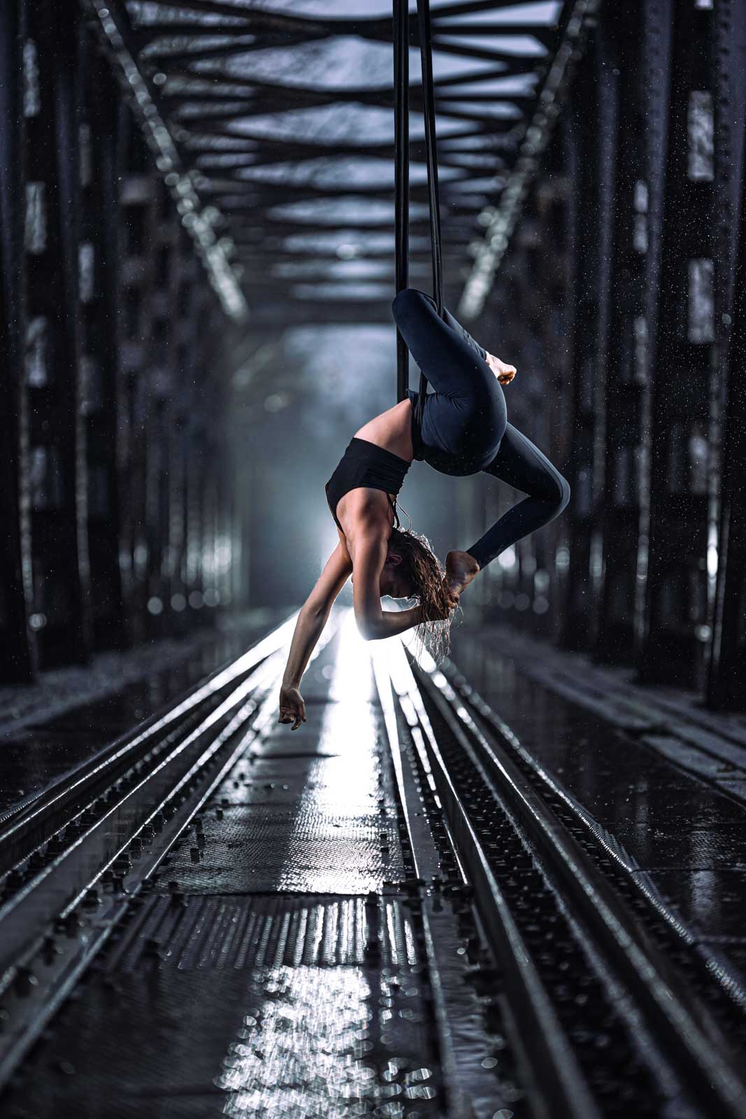 Akrobatin an den Strapaten hängt vor beindruckender Kulisse einer verlassenen Eisenbahnbrücke im Regen.