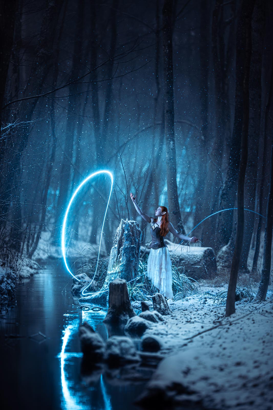 Fantasy / Cosplay Fotoshooting in München: Frau im Elfenkostüm steht nachts im Wald. Es schneit und ein blauer Lichtschweif beleuchtet die Szene.