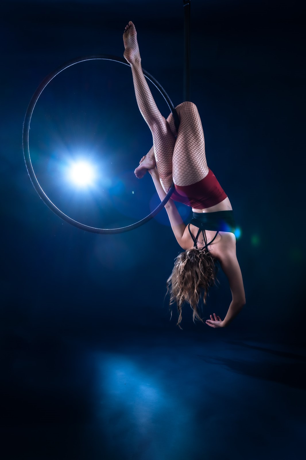 Akrobatik-Fotoshooting im Studio: Frau hängt am überkopf am Aerial Hoop vor dunkelblauem Hintergrund und wird vom Blitzlicht beleuchtet.