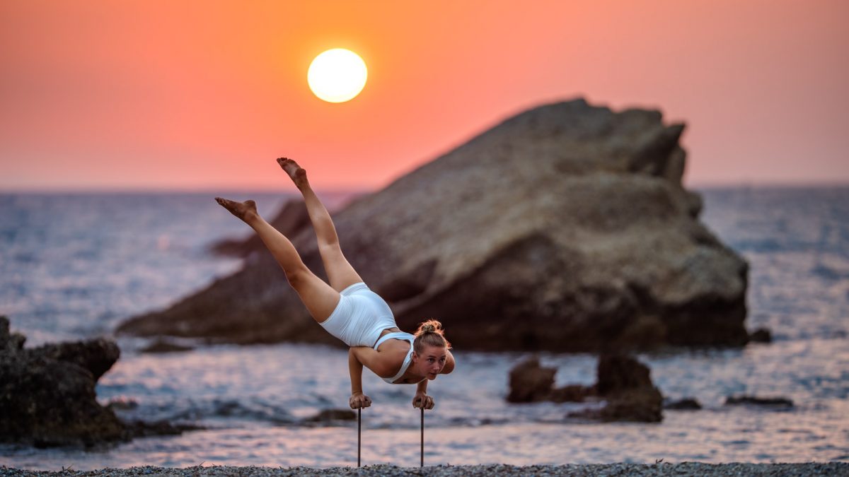 Frau auf Handstand-canes vor Felsen im Meer im Sonnenuntergang.