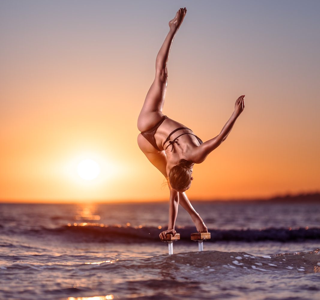 Handstand Fotoshooting: Frau steht im einarmigen Handstand auf Griffen im Meer. Im Hintergrund geht die Sonne unter.