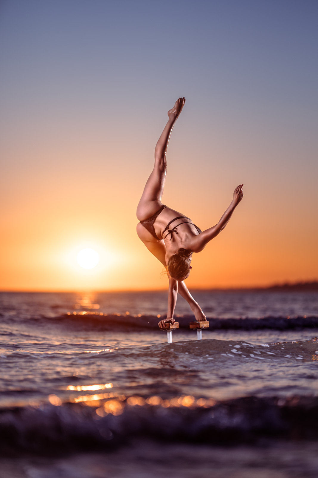 Handstand Fotoshooting: Frau steht im einarmigen Handstand auf Griffen im Meer. Im Hintergrund geht die Sonne unter.