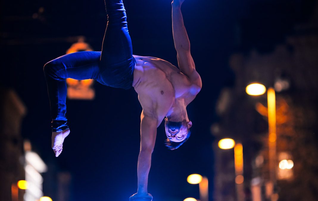 Handstand-Akrobat steht auf einer Hand auf leuchtenden Stäben bei Nacht an einer Straßenkreuzung in der Stadt.
