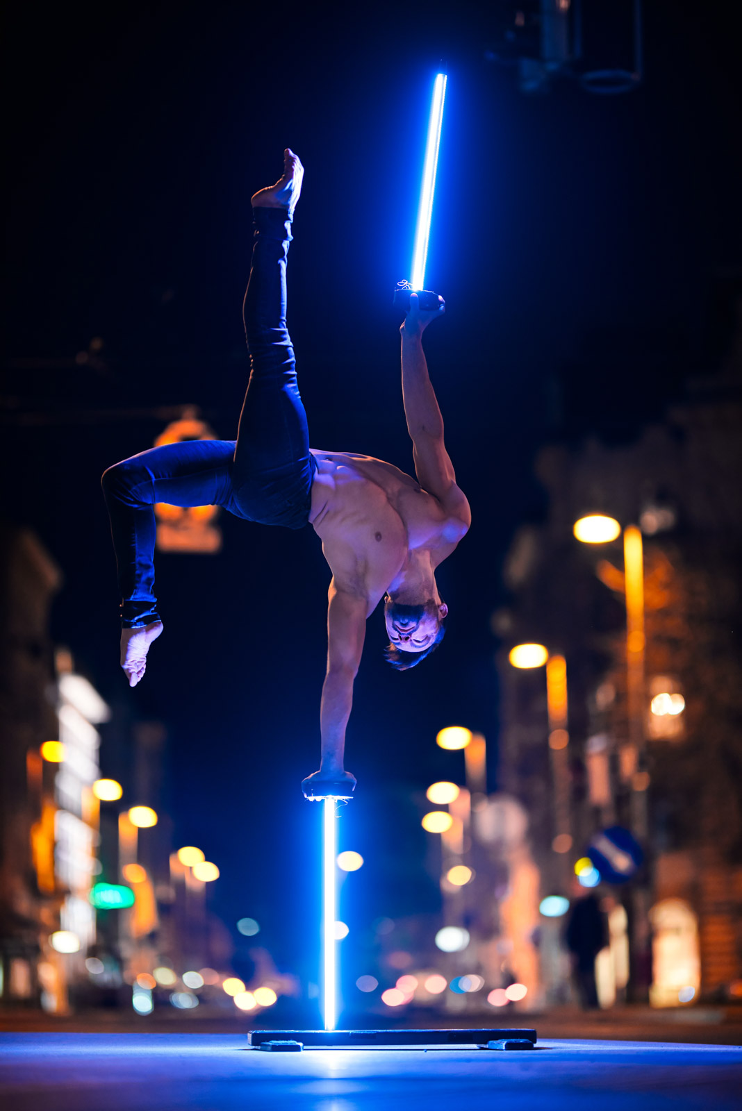 Handstand-Akrobat steht auf einer Hand auf leuchtenden Stäben bei Nacht an einer Straßenkreuzung in der Stadt.