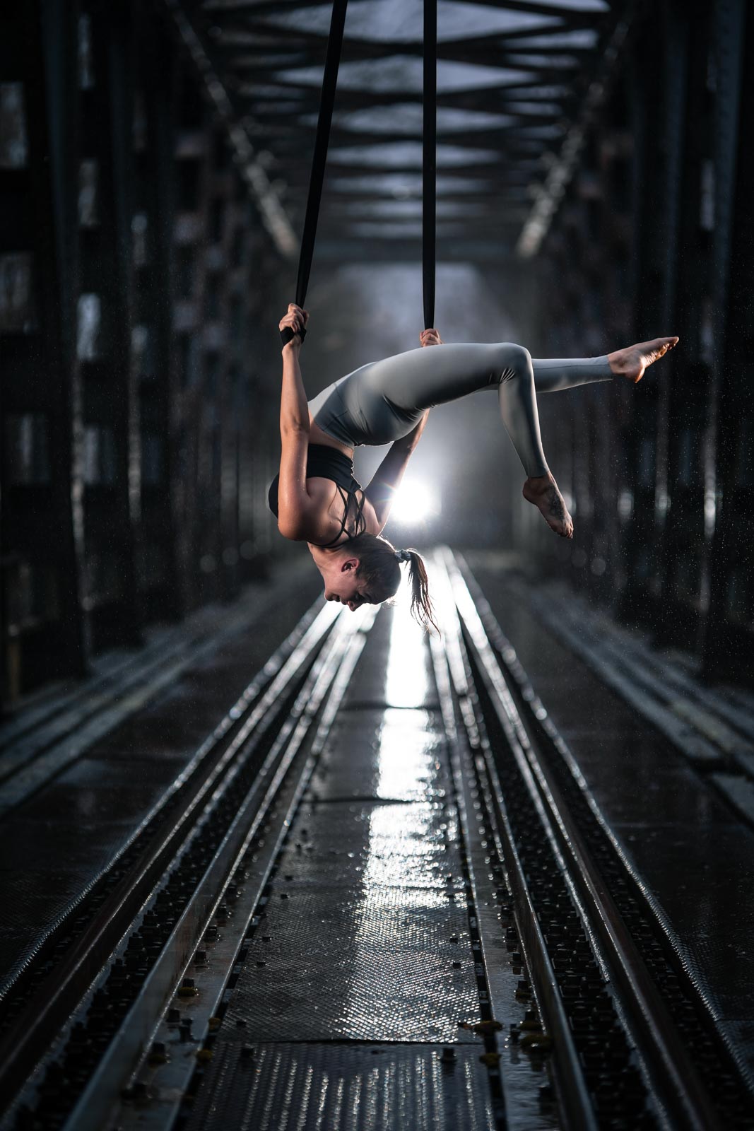 Luftakrobatik Artistin hängt an einer Brücke über verlassenen Bahngleisen im Regen. Blitzlicht sorg für dramatische Beleuchtung.