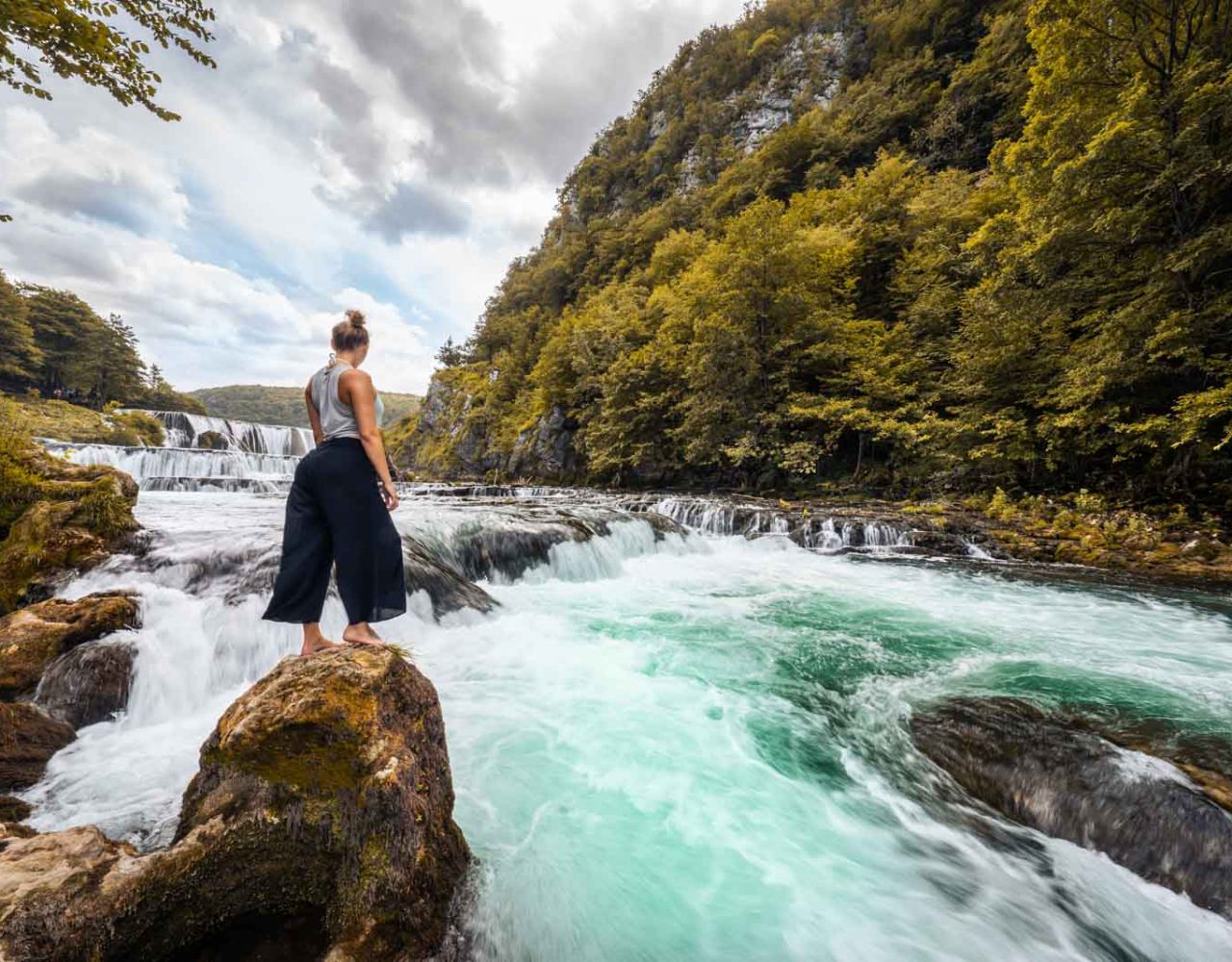 Frau steht am Fluss mit Wasserfällen im Hintergrund.