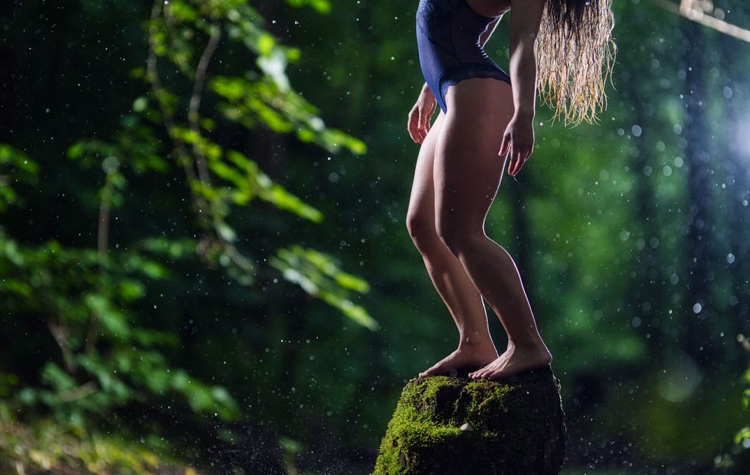 Frau steht bei Nacht im Regen in Dessous auf einem Stein in einem Fluss. Ein Scheinwerfer beleuchtet die Szene.