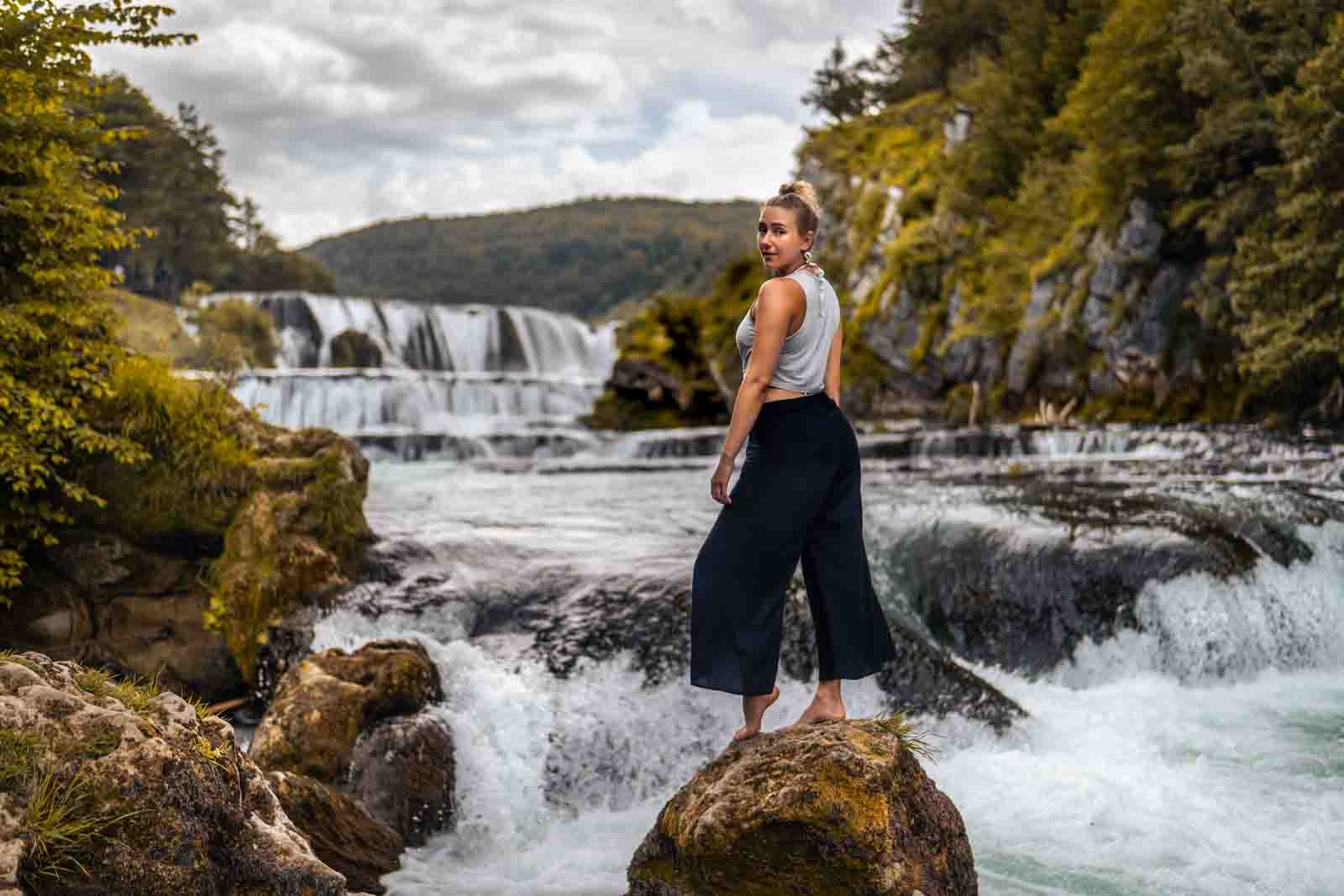 Outdoor-Ganzkörperportrait: Frau steht auf einem Stein im Fluss mit Wasserfall im Hintergrund.