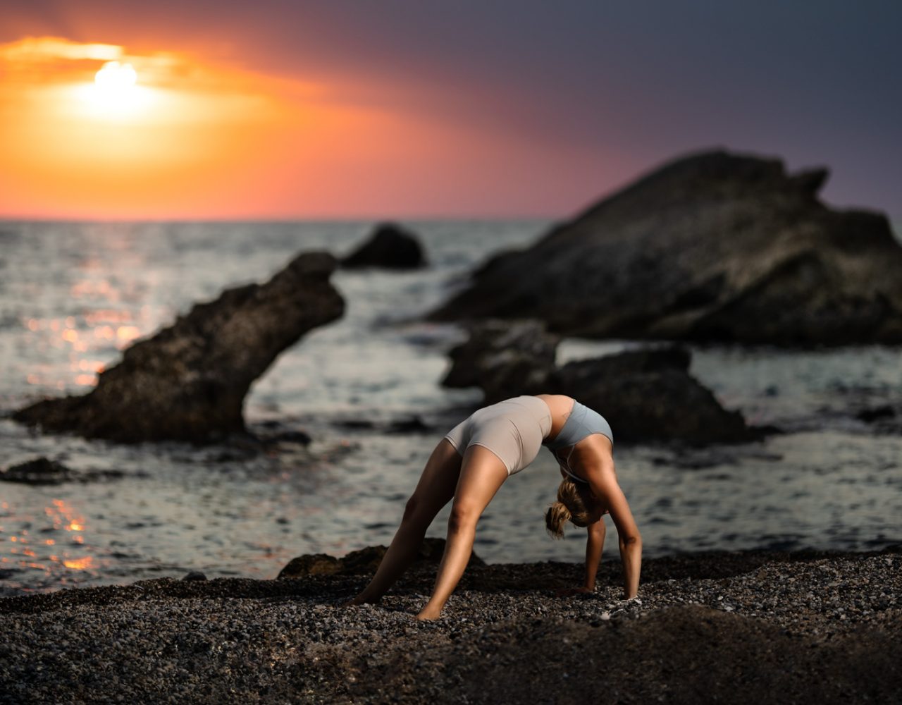 Frau macht eine Bruecke am Strand vor einem Dramatischen Sonnenuntergang. Im Hintergrund befinden sich Felsen im Meer.