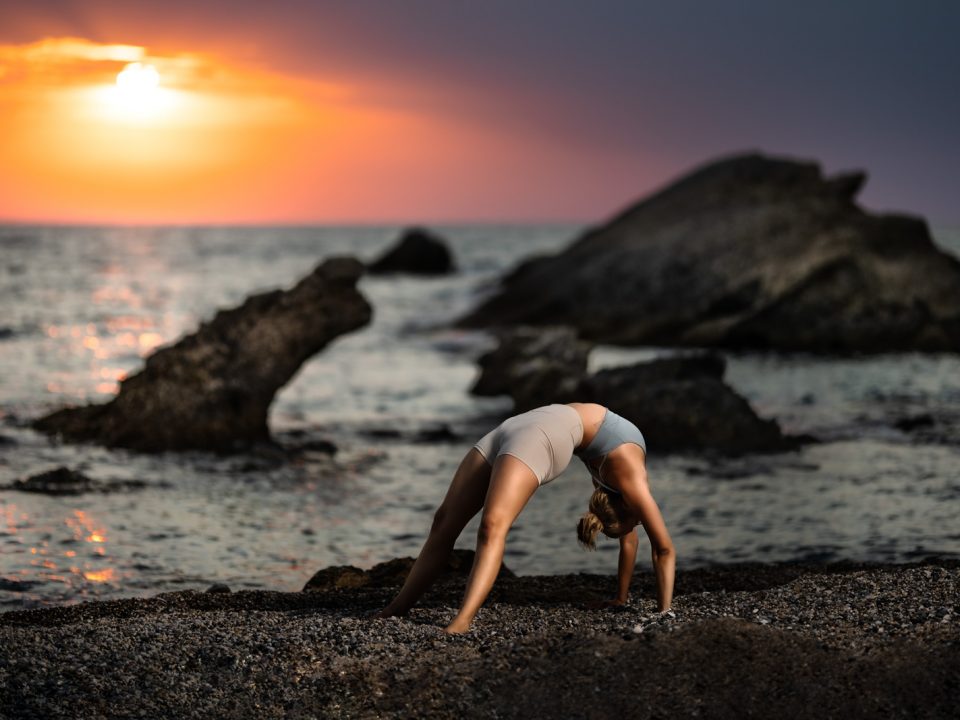 Frau macht eine Bruecke am Strand vor einem Dramatischen Sonnenuntergang. Im Hintergrund befinden sich Felsen im Meer.