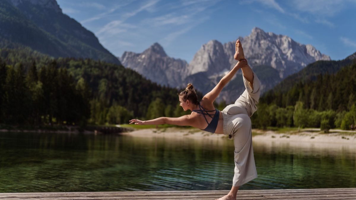 Yoga / Tanz-Fotogshooting: Tänzerin in eleganter Pose auf einem Bein vor schöner Landschaft mit See und Bergen im Hintergrund.