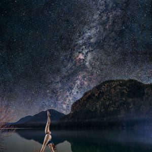 Frau macht einen stehenden Spagat unter dem Nachthimmel vor einem Bergsee.