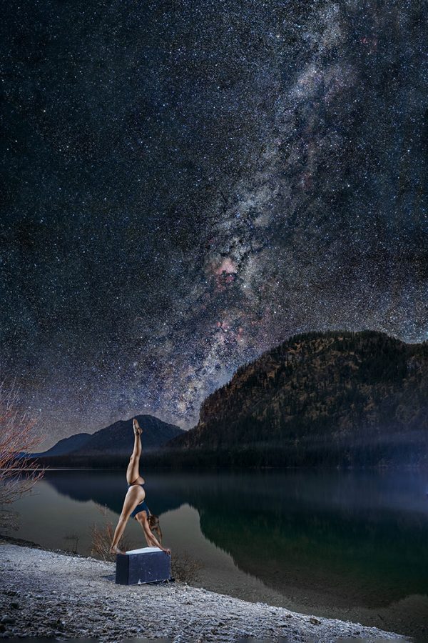 Frau macht einen stehenden Spagat unter dem Nachthimmel vor einem Bergsee.